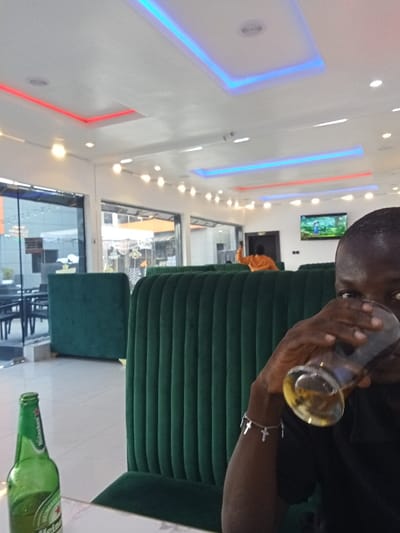 The image shows the interior of a restaurant in Ujevwu, Nigeria (latitude: 5.472, longitude: 5.817). A man in a black shirt is sitting at a booth. He is holding a glass of amber liquid to his lips and appears to be drinking. There is a bottle of beer on the table. The booth is upholstered in dark green fabric. The restaurant has a white ceiling with red and blue light strips. The back wall has a large window, showing a view of the exterior of the restaurant. The glass windows reflect the light coming from outside. In the background, another person is visible standing behind a booth. The background has additional seating in the form of booths. The person behind the booth is wearing an orange shirt. The restaurant has several lights on the ceiling. There are no other people visible in the image.