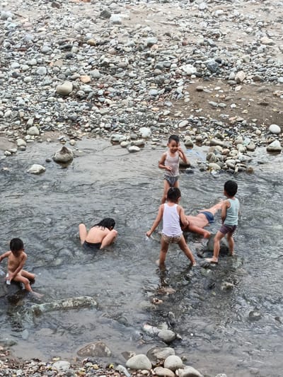 The scene depicts six children playing in a shallow, rocky river in Meurah, Indonesia. The riverbed and banks are covered with numerous smooth, rounded stones and pebbles of varying sizes and muted colors, interspersed with some sand and small bits of debris. The water exhibits ripples from movement and appears generally shallow, allowing the children to stand or sit.
On the far left, a young boy, shirtless and wearing dark bottoms, sits on a partially submerged rock, appearing to hold a small white object. To his right and slightly further into the water, another child with dark hair, wearing dark bottoms, is crouched facing away from the camera. In the center of the frame, an older child or young adult lies supine in the water, wearing dark blue shorts, with their head submerged or obscured. Three other children stand around this prone individual. One young boy in a white tank top and patterned briefs stands to the right of the prone child, looking towards the left, with a hand near his face. Another young girl, facing away from the camera, stands to the left of the prone child, wearing a white tank top and patterned briefs, holding a white object in her right hand. To the far right, a young boy in a light blue tank top and dark briefs stands upright, looking towards the prone child. The overall impression is one of recreational activity in a natural, stony water environment.