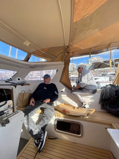 The scene depicts the interior of a boat, likely a yacht or catamaran, docked within a marina in La Grande-Motte, France. An older male, wearing a dark blue long-sleeved shirt, dark grey jeans, a blue baseball cap, and dark athletic shoes with white soles, is seated on a white upholstered bench on the port side of the vessel. Two brown paper bags rest on the bench beside him. Opposite, on the starboard side, an older female with white hair and white-framed sunglasses, dressed in a light grey long-sleeved top, shiny silver-grey pants, and black and white athletic shoes, is seated on a raised white platform. An open, rectangular cardboard pizza box, containing several small, partially consumed pizza remnants, is positioned on a wooden serving board or table between the two individuals. A dark, patterned blanket or fabric is folded on the bench next to the female. The boat's interior features white fiberglass panels, a wooden-slat deck, and an overhead canvas canopy that is partially beige and partially white. Through the boat's forward windows, the background shows other boats docked in the marina and distant multi-story white buildings consistent with the architecture of La Grande-Motte, under a clear blue sky. Bright sunlight illuminates the scene, casting defined shadows inside the boat.