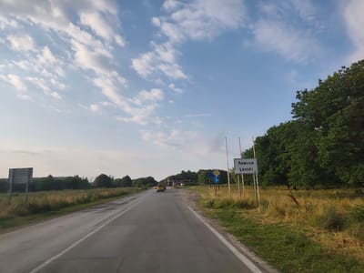 The image depicts a view from a vehicle traveling on a two-lane asphalt road in Levski, Bulgaria. On the right side of the road, there is a white sign with "Левски" in Cyrillic and "Levski" in Latin script, indicating the entrance to the town. Adjacent to the sign is a dense green tree line. The road itself shows signs of wear, with some cracks and patches. On the left side of the road, there is a grassy verge and a large, empty rectangular billboard. In the distance, a yellow car is visible on the road. The sky is blue with scattered white clouds. The overall scene suggests a rural or semi-rural approach to the town of Levski.