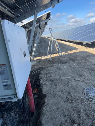 The image displays a ground-mounted solar photovoltaic power plant located in Polkovnik Lambrinovo, Bulgaria. The foreground on the left features a white electrical enclosure, partially open, with a visible "HUAWEI" label. From the bottom of this enclosure, multiple black corrugated electrical conduits and one prominent red corrugated conduit extend downwards into an exposed trench. This trench, filled with additional conduits and loose soil, runs horizontally across the midground. To the right of the trench, an open silver aluminum A-frame step ladder stands on the dry, uneven dirt ground. The background reveals extensive rows of blue solar panels mounted on grey metal support structures, extending into the distance under a partly cloudy blue sky. The overall ground surface is dry, dusty soil with scattered debris. No individuals are present in the scene.