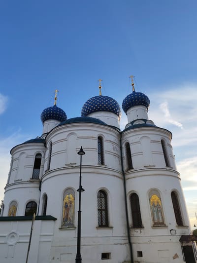 The image shows the Church of the Assumption of the Blessed Virgin Mary in the village of Chelganovo, Russia. The church features white walls and a dark green roof, topped with three prominent blue domes adorned with gold stars. Each dome is crowned with a golden cross. The building has several arched windows with dark frames and decorative mosaic icons set into the walls. A black lamppost stands to the right of the church. The sky above is clear blue with a few wispy clouds, and a hint of sunlight illuminates the scene. The church appears to be an Orthodox Christian place of worship.