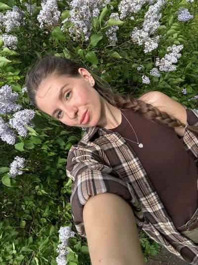 A young woman lies in a field of lilac flowers in Борщевка Малая, Russia. She has light skin, blue eyes, and her brown hair is braided. She wears a brown ribbed tank top with a silver heart-shaped necklace and a brown plaid flannel shirt.