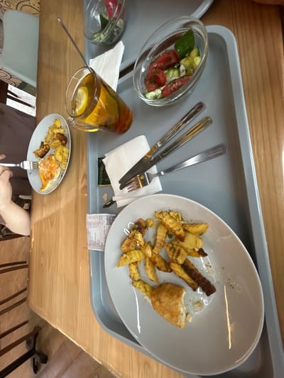 The image depicts a high-angle, full-frame shot of a gray food tray resting on a wooden table. The tray contains a meal that appears to be partially consumed. On the tray, there are two white plates, one with a piece of fried fish and a portion of french fries, and another smaller white plate with french fries. A clear glass mug filled with a golden-colored beverage and a slice of lemon is positioned on the left side of the tray. Adjacent to the mug is a small glass bowl containing a salad of chopped tomatoes and cucumbers. Cutlery, including forks and knives, is laid out on white napkins. A crumpled receipt is also visible on the tray. The background is slightly blurred, showing glimpses of a patterned surface and furniture, suggesting an indoor dining setting. The overall scene suggests a casual meal experience. The prompt's request to include the location in the city of Inzer, Russia, cannot be fulfilled as there are no visual cues in the image to support this information.