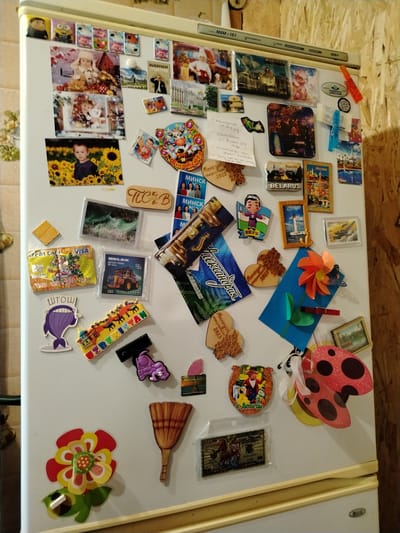 The image shows a white refrigerator door covered with various magnets and photographs. The magnets are in diverse shapes and sizes, featuring colorful illustrations and text. Some magnets appear to be souvenir magnets from different locations, including what looks like a magnet with "BELARUS" written on it and another that says "UZBEKISTAN". There are also personal photographs attached, depicting children and family scenes. The overall impression is one of a decorated, personal refrigerator. The location in the city of Рудзенск, Belarus, is not explicitly identifiable from the image itself, as it depicts the interior of a home appliance.