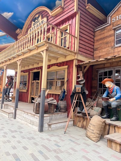 The image depicts a western-themed attraction, likely a restaurant or shop, in the city of Collendoorn, Netherlands. In the foreground, a paved area with a pattern of gray and reddish-brown bricks leads to the entrance of the establishment. The building itself is constructed with weathered red and brown wood siding and features a prominent second-story balcony with a wooden railing.

On the left side of the entrance, a life-sized mannequin of a cowboy stands on the wooden porch, wearing a brown hat, jacket, and jeans. Beside him, a wooden bench is visible. The main entrance has double swinging doors with wooden shutters.

To the right of the entrance, a second mannequin depicts an elderly cowboy seated on a wooden step. He wears a brown hat, a plaid scarf, and blue overalls, and holds a bowl and a cup. In front of him, there are several burlap sacks, one of which is labeled "COFFEE-14/LIP" and "WEIGHT: 50 KGS". Various other western-themed props are scattered around, including boots hanging from a beam, a vintage camera on a tripod, a saddle on a wooden crate, and barrels. A blue acoustic guitar leans against a barrel.

The facade of the building above the balcony displays a sign that reads "BOOTHLILL SALOON". The sky above is a bright blue with white clouds, appearing to be artificial, suggesting an indoor or partially covered space designed to simulate an outdoor environment. The overall scene is a static, staged display designed to evoke a Wild West atmosphere.