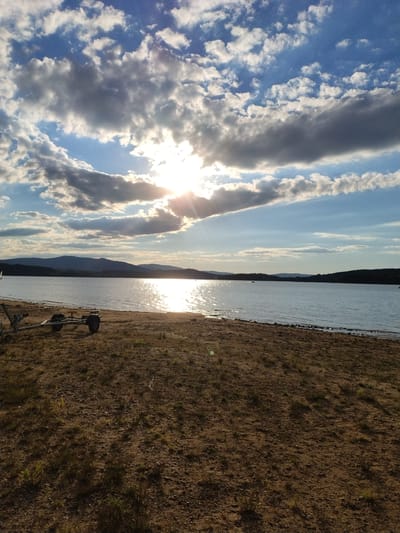 The image depicts a serene lake scene at sunset. The sun, partially obscured by dramatic clouds, casts a brilliant reflection on the calm water. In the foreground, a sandy shore is visible, with sparse dry grass and a boat trailer with two wheels. The background features a chain of hills and a forest. The sky is a mix of blue and grey clouds, with rays of sunlight breaking through. The overall atmosphere is tranquil and picturesque. The location is identified as Radslav, Czechia.