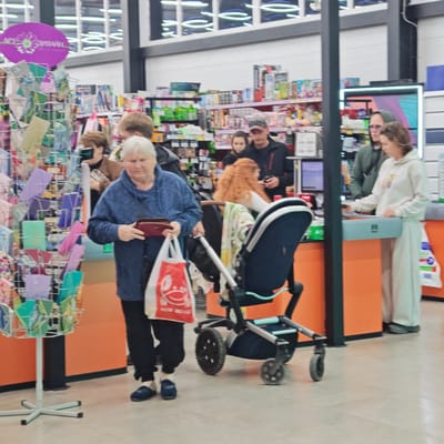 The image captures an indoor scene within a store, likely a supermarket or general merchandise outlet, focusing on a checkout area. Several people are present, engaged in shopping activities.
In the foreground, an elderly woman with white hair, wearing a blue sweater and dark pants, holds a red wallet in one hand and a white shopping bag with a red design in the other. The text "ВСЁ ДЛЯ ДОМА" (All for Home) is visible on the bag, along with a red smiley character. To her right, a dark-colored baby stroller with light blue accents and a patterned blanket draped over it stands next to her. Behind the stroller, a woman with long red hair is visible, and further back, a man in a dark jacket and baseball cap.
To the right side of the frame, a young woman in a white hooded sweatshirt and light-colored pants is standing at a checkout counter, seemingly making a purchase. A man in a green hooded jacket and glasses stands behind her. The checkout counters feature orange bases and light blue top panels, one of which displays a "回" symbol. Computer monitors and barcode scanners are visible on the counters.
The background shows numerous shelves stocked with various products, indicating a retail environment. A metal display rack with colorful greeting cards or small stationery items is positioned to the left. Above this display, a purple oval sign reads "АРТ СУВЕНІР" (Art Souvenir) with a daisy graphic, suggesting a gift or craft section. The store has a modern appearance with white walls, black structural pillars, and overhead fluorescent lighting that reflects in what appear to be high windows. The floor is covered with light-colored tiles. The Cyrillic text visible on signs aligns with the provided location context of Ukraine. The overall atmosphere is that of a busy but orderly retail environment.