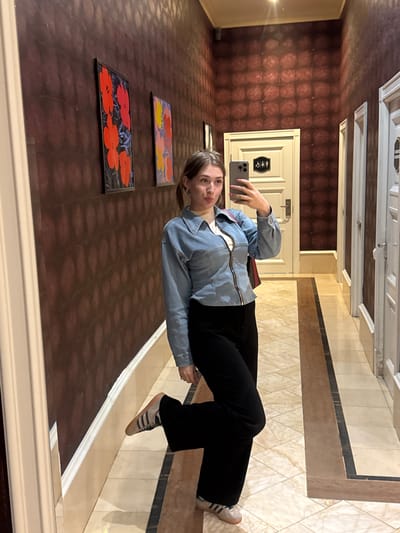 A young woman stands in a hallway and takes a mirror selfie. She is wearing a light blue denim jacket with a zipper down the front, a white shirt underneath, and black pants. She has on beige Adidas sneakers. She is holding a smartphone in her right hand, angled to capture her reflection. The hallway has dark reddish-brown textured wallpaper and a tiled floor with a patterned border. Two framed paintings of flowers hang on the wall to the left of the woman. A white door with a sign indicating restrooms is visible behind her. The lighting in the hallway is warm. The location is the city of Shkotovo, Russia.
