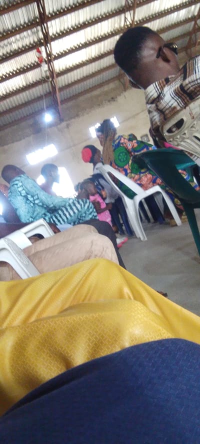 The image shows an interior scene, likely a community hall or church in Ibo, Nigeria. In the foreground, a person wearing a yellow garment is seated, with a portion of a blue garment visible below. Further back, several individuals are seated on white plastic chairs. One person is wearing a patterned blue and green dress, and another person has a red head covering. The ceiling is made of corrugated metal, supported by a metal structure. Natural light filters in from rectangular openings in the ceiling. The atmosphere appears to be a gathering or service.
