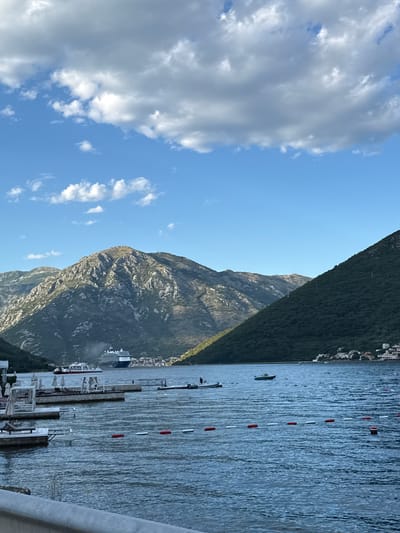 This is a vertical, eye-level, full shot of a body of water with mountains in the background. The water is dark blue and choppy, with a few buoys and boats floating on it. In the background, there are large, rugged mountains covered in greenery. A cruise ship and a ferry are visible on the water, and some buildings are nestled into the mountainside. The sky is blue with scattered white clouds. A pier with lounge chairs and umbrellas is in the foreground on the left. The scene appears to be in a bay or fjord, with mountainous terrain and water. The location is Jošice, Montenegro.