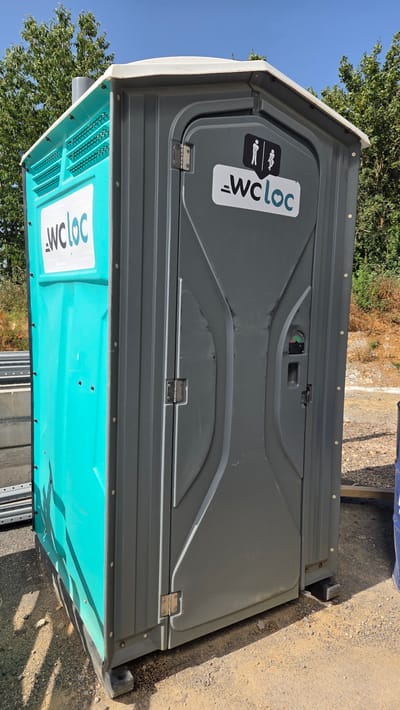 This is a vertical, eye-level, outdoor photograph of a portable toilet. The toilet is dark grey on top with a turquoise blue base. It is situated on a gravelly surface, with dry brush and a large green tree visible in the background. The sky is clear and blue.

There are two logos visible on the toilet. The upper logo, on the grey portion of the toilet door, is a black and white stylized image of a person, accompanied by the text "WCloc" in white letters with a light blue outline. The lower logo, on the turquoise base, is a white version of the "WCloc" text.

The toilet door has a silver latch with a green and red indicator. The door is held closed by hinges on the left side. The base of the toilet has a vented section with horizontal slats.

The lighting suggests it is daytime, likely sunny, given the clear sky and shadows. The overall scene appears to be a rural or outdoor event setting. There are no people actively using the toilet. The image is crisp and clear, focusing on the details of the portable toilet. The provided location context is Moulins-sur-Orne, France.