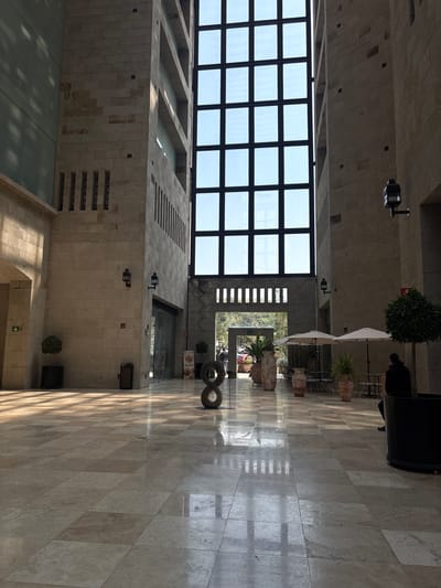 The image shows an interior shot of a building in Ciudad López Mateos, Mexico. The architecture features a high ceiling with a large grid of windows that allow natural light to illuminate the interior. The floor is tiled and reflects some of the light.
To the right, there are potted plants and seating under white umbrellas, suggesting a cafe or lounge area. A person is seated at one of the tables. An art piece resembling a figure eight is visible in the foreground.
The walls are made of light-colored stone blocks with recessed designs. Security cameras are mounted on the upper portion of the walls. A sign is visible near the entrance to a store or restaurant.
