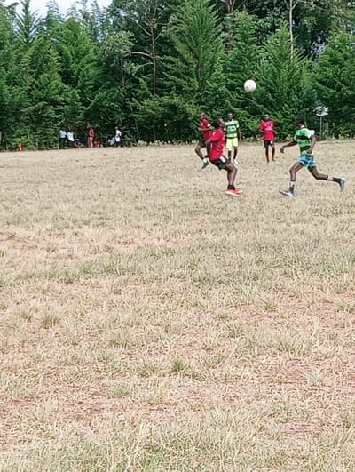 An outdoor football match is in progress on a dry, grassy field in Lwandeti ward, Kenya. A male player wearing a red jersey, dark shorts, and orange footwear is captured mid-air, having just kicked a white football with his right foot. The football is visible above and slightly to his right. Another male player in a green and black striped jersey, patterned shorts, and dark footwear is running towards the right in the mid-ground. Additional players in red and green/black jerseys are positioned across the field, indicating active play between two teams. A dense line of tall green trees forms the background, with several indistinct individuals visible along the tree line to the left. The field itself consists of light brown and green sparse grass with visible patches of earth.