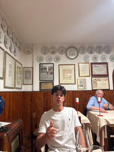 In the city of San Biagio di Callalta, Italy, at what appears to be a restaurant or dining establishment, two individuals are seated at tables. The foreground features a young man with dark hair, wearing a white t-shirt with a blue embroidered logo, facing the camera. He is gesturing with his right hand. Behind him, to the right, an older man with gray hair, dressed in a blue button-down shirt, is seated. Both individuals are positioned at tables covered with light-colored tablecloths, each adorned with a white candle in a red holder. The background reveals a wall decorated with numerous framed plates and documents, along with dark wooden paneling. A wooden cabinet or buffet is visible to the left. The lighting suggests an indoor setting, likely in the evening.