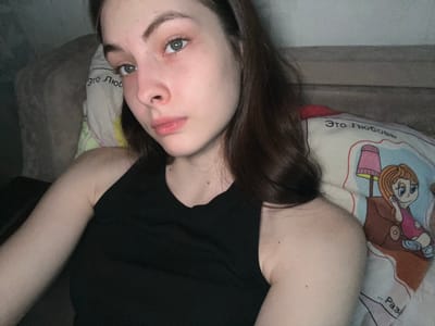 A woman is lying down, possibly on a bed, with a pillow under her head. She has dark hair and fair skin. She is wearing a black top. The pillow has a cartoon illustration and the words "Это любовь" (This is love) written on it in Russian. The background is partially visible, showing what appears to be a wall and a sofa or bed frame. There is not enough information to identify a location in Shcheglovo, Russia.