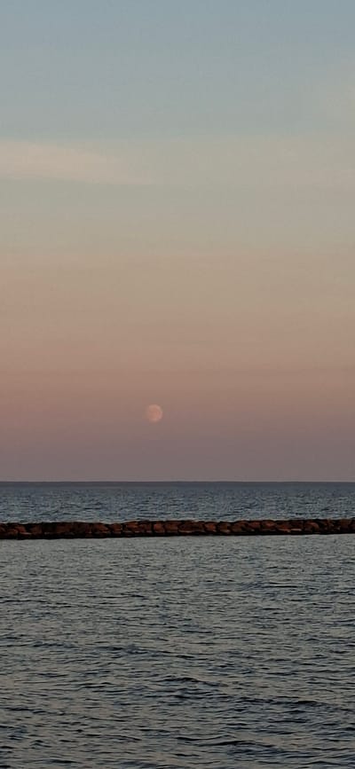 The image shows a view of the Baltic Sea from Pärnu, Estonia, at dusk. The sky is a gradient of pale blue and pink, with the moon visible as a faint, pinkish circle in the upper portion of the frame. A stone breakwater stretches horizontally across the middle of the image, with the dark blue water of the sea extending below it. The overall impression is one of tranquility and natural beauty.