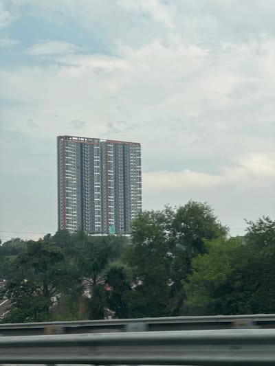 The image shows a modern, multi-story residential building with a grey facade and orange trim. The building is tall and rectangular, with many windows arranged in a grid pattern. It appears to be under construction or newly built, with scaffolding visible on one side. The building is surrounded by lush green trees, suggesting a natural or semi-urban setting. The sky is overcast with clouds, indicating a possibly humid or rainy day. The image is taken from a slightly elevated perspective, possibly from a moving vehicle, as evidenced by the blurred vertical metallic structure on the right side of the frame. There is no visible text in the image. The scene depicts a slice of urban landscape in a tropical or subtropical region, likely in Malaysia given the location context. The overall mood is neutral, focusing on the architectural subject and its environment.