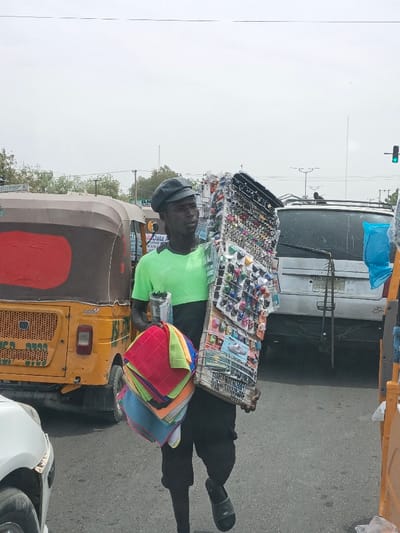 The scene depicts a daytime urban street in Hotoro, Nigeria. A dark-skinned male street vendor, wearing a black newsboy-style cap, a lime green and dark two-tone t-shirt, and dark shorts or trousers with black slide sandals, is positioned centrally, walking forward. He carries two distinct sets of merchandise: in his left arm, a stack of brightly colored, folded cloths or towels (red, lime green, blue, yellow, grey); and in his right arm, a large, rectangular display board showcasing numerous small items such as sunglasses, keychains, and other trinkets arranged on hooks.
In the immediate background to the vendor's left is the rear of a yellow and brown tricycle auto-rickshaw, displaying a red rectangular window and white lettering including "KANO STATE." Further back, behind the vendor, a silver or light-colored SUV or minivan is visible, partially obscured. A small portion of a white vehicle is present in the lower-left foreground. The road surface is asphalt. Utility poles, wires, and a green traffic light are visible against an overcast sky. Distant buildings and sparse vegetation line the horizon.