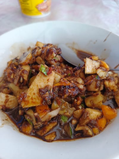 A close-up, top-down view depicts a white ceramic bowl filled with a dark-sauced fruit and vegetable salad, consistent with Indonesian Rojak. The contents are primarily diced, exhibiting varied textures and colors: light beige squares, vibrant orange-yellow wedges, and darker green fragments, all heavily coated in a thick, glossy, dark brown to black sauce. This sauce shows specks of lighter material, possibly ground peanuts or chili. A silver spoon is partially visible within the upper right section of the bowl, immersed among the ingredients and sauce. The bowl rests on a light, indistinct surface. In the blurred upper-left background, a yellow cylindrical object with red and white branding, resembling a packaged food or beverage container, is partially visible. No people are present. The specific location in Meurah, Indonesia, is not visually ascertainable from the image.