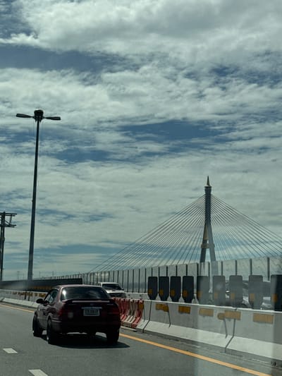 The image shows a view from a vehicle on a highway in Song Khanong Subdistrict, Thailand. A dark red sedan is in the foreground, driving in the right lane. Behind it, another car is visible. The highway has a concrete barrier on the right side, with yellow and orange traffic cones or markers. In the background, a large cable-stayed bridge dominates the scene, with its tall pylon and numerous suspension cables. A tall street lamp with multiple lights is on the left side of the highway. The sky is cloudy with patches of blue visible.