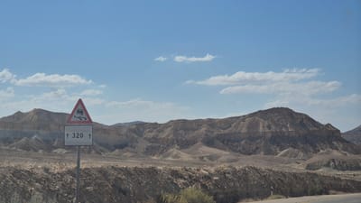 This image depicts an arid desert landscape within the Ramat Negev Regional Council, Israel, under a clear, light blue sky dotted with scattered white cumulus clouds. In the foreground, slightly to the left of center, a metal pole supports two road signs. The upper sign is a standard red-bordered warning triangle featuring a black pictogram of a car skidding, indicating a slippery or hazardous road condition. Below it, a white rectangular sign displays the black text "↑ 320 ↑", likely representing a road marker or elevation. The immediate foreground consists of rocky, barren terrain with sparse, low-lying scrub brush. Extending across the midground and into the background are numerous rocky, stratified mountains and hills, characteristic of a desert environment, exhibiting various shades of brown, tan, and grey due to their geological composition and erosion patterns. No people, animals, or vehicles are visible within the frame.