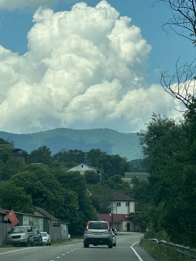 The image depicts a scenic view of a rural road in the vicinity of Grecheskii, Russia, under a dramatic sky filled with large, white cumulus clouds. The road, a paved asphalt surface with a white dashed line marking its center, curves gently to the right. On the left side of the road, several vehicles are parked, including a silver SUV, a dark SUV, and a white sedan. A red flag is visible on a pole outside a building with a grey fence. Various structures, including houses with white and yellow facades and dark roofs, are nestled amongst lush green trees on the left and right sides of the road. In the background, rolling green hills rise, partially obscured by trees. The overall impression is of a tranquil, somewhat isolated, natural landscape within a populated area.