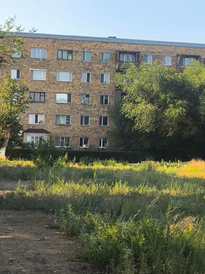 The image shows a multi-story apartment building constructed from light brown bricks. The building has numerous windows, some with white frames and curtains, and others with dark frames. Several balconies with wooden railings are visible on the upper floors. In front of the building, there is dense green foliage, including trees and bushes. A patch of dry, sunlit grass is in the foreground. The sky above is clear and blue. The scene appears to be a typical residential area in the city of Abay, Kazakhstan, under bright daylight.