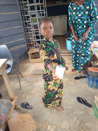 In Iseyin, Nigeria, a young girl, appearing to be around 6-8 years old, stands on a concrete floor in front of a store. She is wearing a black, green, and yellow patterned dress and holds a white box in her right hand. To her right, a woman in a blue, green, and white patterned dress stands, also holding a small object in her hand. The background reveals the interior of a store with shelves stocked with various items and a white refrigerator. The area outside the store appears to be a marketplace, with a metal gate, wooden stools, buckets, and discarded flip-flops visible. The overall scene suggests a typical day at a local market in Iseyin.