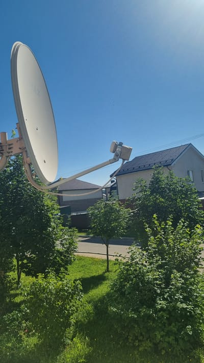 In the town of Verzilovo, Russia, on a clear, sunny day, a satellite dish is mounted on a structure, angled towards the bright blue sky. The dish, a light color, has the word "MAX" visible on its surface. It is supported by a metal arm extending from a base. Behind the dish, lush green trees and bushes create a natural foreground. In the background, residential houses with dark roofs are visible, along with a paved road and more green spaces. The overall scene depicts a typical suburban or rural environment on a pleasant day.