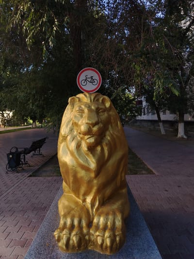 A golden lion statue sits on a stone pedestal in the foreground. Above the statue, a circular traffic sign with a red border and a bicycle symbol crossed out is visible, indicating no cycling is allowed. The statue is positioned on a paved walkway made of interlocking bricks, with a black bench to the left. Lush green trees with dense foliage form a canopy overhead, casting shadows on the scene. Beyond the walkway, another paved path leads towards buildings in the background. The setting appears to be a park or public area in the city of Baikonur, Kazakhstan.