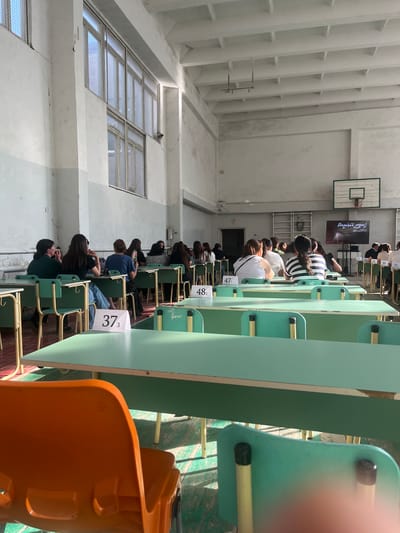 This is an interior shot taken within a school gymnasium or hall in Gladzor, Armenia, likely during a formal event or examination. The scene is dominated by rows of light green student desks and chairs, arranged in neat columns. Several young people are seated at the desks, facing away from the camera, their backs turned as if engaged in a test or lecture. The students are dressed casually, with many wearing striped shirts or plain t-shirts.

At the far end of the room, a basketball hoop and backboard are visible mounted on the wall, suggesting the multipurpose nature of the space. A large screen is also present, displaying text that appears to be a title or announcement. The lighting suggests it's daytime, with natural light filtering in through large, barred windows on the left side of the image. The overall atmosphere seems quiet and focused, typical of an academic setting.

Visible text includes numerical labels on the desks such as "373", "471", and "482", likely indicating student seating assignments. There are also partial words visible, such as "RISMAS" and "Amit ymh mbunt," possibly part of a longer inscription or branding. The orange plastic chair in the foreground and the metal supports of the desks provide some textural contrast to the predominantly white and green color palette of the room.
