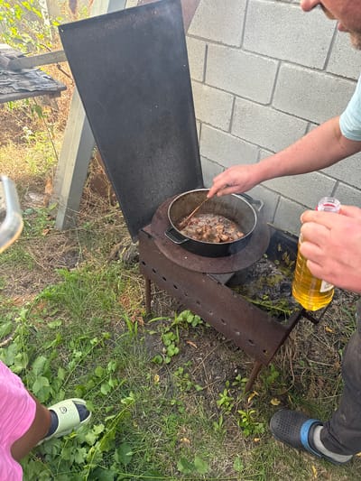 A person's arm is seen pouring yellow liquid from a plastic bottle into a metal pot that is placed on a makeshift metal stove. The pot contains chunks of meat that are being cooked in the liquid. Another person's arm is visible stirring the contents of the pot with a wooden spoon. The scene appears to be outdoors, with a paved area on the left and grass on the right. The setting suggests a casual outdoor cooking activity, possibly a barbecue or a gathering. The lighting indicates it is daytime. The focus is on the food preparation process.