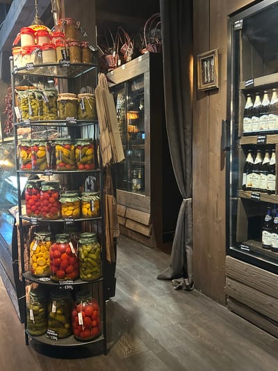 The image depicts a retail display of preserved food items, predominantly in glass jars, within a rustic-themed establishment. The display shelves, made of black metal, are laden with jars containing pickles, tomatoes, and other pickled vegetables. Some jars have handwritten labels indicating the contents and prices.
To the right of the pickled goods, a refrigerated display case showcases numerous bottles of beer, primarily labeled "KUMBACHER". These bottles are arranged on shelves within the illuminated case. The overall environment appears to be a shop or a market stall.
A dark brown curtain hangs on the left side of the frame, partially obscuring a darker interior space. The walls are constructed of dark wood planks. A framed picture containing cutlery is hung on the wooden wall. The flooring is a dark wood laminate. The setting suggests a local market or specialty food store in Новоглаголево, Russia, focusing on artisanal preserved foods and beverages. No people are visible in the image.