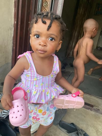 A young girl with braided hair and gold earrings holds up pink Crocs. She wears a pink and white striped dress with lemon patterns and floral shorts. In the background, a naked toddler stands on what appears to be a porch. Several pairs of shoes are scattered on the ground near the two children. The lighting is bright, with natural light illuminating the scene.