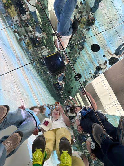 The image shows a group of people in a mirrored installation. The perspective is from the ground, looking upwards. The reflections create a dizzying effect, multiplying the people and the environment.

The subjects are dressed in casual clothing, including jeans, T-shirts, and sneakers. Some are holding phones and taking photos. The environment appears to be an art installation with mirrored walls and ceiling, set on a floor with a light blue and white pattern, potentially simulating water or clouds.

The location is potentially within a building or structure in Haría, Spain.
