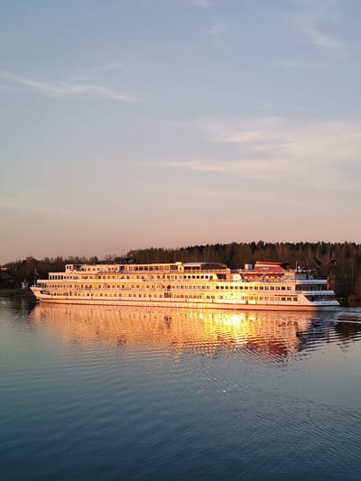 A long white river cruise ship is on the water near 3-Y Uchastok, Russia. The ship has multiple decks with numerous windows. The setting sun reflects strongly on the ship's surface and in the water, creating a golden glow. The sky is a mix of light blue and soft pink. Trees line the bank in the background.