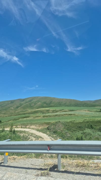 This is a wide-angle, slightly blurred photograph taken from a vehicle moving along a road. The image captures a rural landscape under a bright blue sky with wispy white clouds. In the foreground, a metal guardrail with reflective markers runs horizontally across the frame. Beyond the guardrail, a dirt road curves into the distance, bordered by green fields and rolling hills. The hills in the background are covered in vegetation, with variations in texture and color suggesting different types of plant life. The overall impression is of a serene, undeveloped natural environment. The location is identified as Berezovka, Kazakhstan.