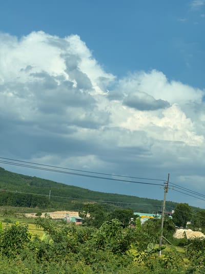 The image captures a landscape in Đăk La, Vietnam, under a partly cloudy sky. A prominent feature is the mountainous terrain covered in dense green vegetation. An electric pole with power lines runs through the scene. A building with a blue roof is visible amidst the foliage. In the foreground, there is more vegetation, possibly bushes or trees. The sky shows a mix of blue and white, with large cumulus clouds dominating the upper left portion of the image.