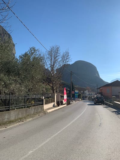 This media file captures a sunny, clear day on a road in Kastraki, Greece, with a massive mountain dominating the background.
The paved road, featuring a white line, curves slightly as it recedes from the lower right foreground towards the upper left background. On the left side of the road, a low concrete wall fronts a black metal fence, behind which lush olive trees with silvery-green foliage and a large, bare deciduous tree are visible. A utility pole with wires runs across the mid-ground.
On the right side of the road, buildings line the street, possibly houses or small businesses. A dark-colored car, appearing to be a hatchback or small SUV, is parked or moving slowly on this side. Several commercial signs are displayed along this stretch. A prominent red banner or awning, oriented vertically, features white text, clearly reading "SOUVENIRS" at the top. Below this, there's more text that is less discernible. Further down the road, a blue and yellow sign, resembling that of a petrol station or convenience store, displays numbers like "1.55" and "1.65," possibly indicating fuel prices.
The most striking feature of the scene is the imposing, greyish-brown mountain, or large rock formation, which rises steeply in the background under a bright, cloudless blue sky. The strong sunlight suggests it is mid-day or early afternoon. The overall setting is semi-urban or rural, characteristic of a village road in a mountainous region like Kastraki, at the foot of Meteora.