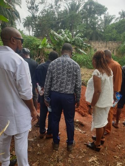 The image shows a group of people standing outdoors, likely at a burial or memorial service based on the somber atmosphere. The ground is reddish soil. The background features dense vegetation including palm trees, suggesting a tropical environment. The location is the city of Nkwerre, Nigeria.

The people are dressed in a variety of clothing. A woman in a short white dress with a lace overlay and braided hair is visible. A man in an orange shirt is next to her. Several men in darker-colored clothing (including patterned shirts) are positioned behind them. One person is wearing a white outfit and sunglasses. They are all looking down at something. Some are holding objects such as a water bottle.

The scene indicates a gathering of people paying respects, possibly at a gravesite.