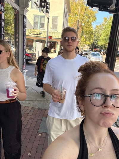 This image captures a street scene on a sunny day in New Hope, Pennsylvania, specifically identifiable by a "Dunkin' Donuts" storefront located across the street.

In the immediate foreground, to the right, a person with red hair, wearing black-rimmed glasses, gold hoop earrings, and two delicate gold necklaces, looks directly at the camera with a slight pout. Behind and slightly to their left, a male with short blonde hair and aviator sunglasses walks forward, holding a clear plastic cup with a straw containing an iced coffee. To the left of the male, a blonde-haired female in dark sunglasses and a light-colored tank top walks, holding a clear plastic cup with a straw containing a purple beverage. Further back on the sidewalk, a person with dark hair, visible tattoos on their left arm, and a black t-shirt with a "PIERCE THE VEIL" graphic walks.

The background shows a moderately active street with pedestrians and several vehicles, including a silver SUV. Multi-story buildings with traditional architecture line the street; one building clearly displays a "DUNKIN' DONUTS" sign above a green awning. A vertical traffic light fixture shows the bottom light illuminated red. Trees in the background exhibit yellow and green foliage, suggesting late summer or early autumn. The immediate ground is a red brick sidewalk. A black banner with white text, partially visible on the left, reads "HAPPY HALL", suggesting seasonal decor.