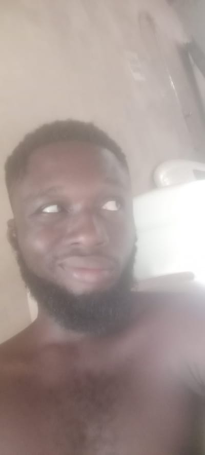 A close-up, slightly blurry image shows the upper torso and head of a dark-skinned man with a full, dark beard. His hair is short and dark. He is looking slightly to his right. His shoulders are bare. The background is a blurred, light-colored wall, with a partially visible white object in the upper right corner that is probably a bathroom fixture. There is no indication of the location or that it is in Bende, Nigeria.