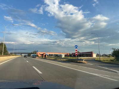 The image shows a highway leading towards buildings with signs that read "AGRO MARKET" and what appears to be a bus stop. There are traffic signs indicating a change in lane direction and a speed limit of 50. The sky is filled with dramatic clouds. The perspective suggests the photo is taken from a vehicle traveling on the highway. The location is identified as Kareli, Georgia.