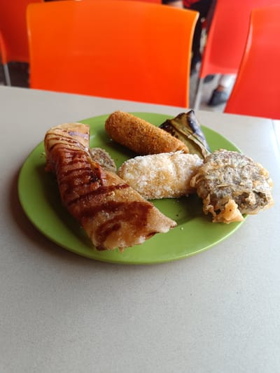 A close-up, eye-level shot presents a vibrant green plate filled with an assortment of fried snacks, resting on a light grey table. In the foreground, a golden-brown, rolled snack, possibly a banana fritter or sweet pancake, is prominently featured, adorned with a generous drizzle of dark chocolate sauce. To its right, a breaded, cylindrical item, golden-brown and crispy, suggests a croquette or similar fried snack. Adjacent to this, a white, sugar-coated fried item with a somewhat irregular shape is visible, likely a sweet fritter. Further right on the plate, a darkly fried, textured, and irregular-shaped item stands out, possibly a savory fritter or fried mushroom. A dark, elongated object, potentially a grilled banana leaf wrap, is partially seen behind the cylindrical snack.

The background is softly blurred, showcasing bright orange and red chairs, indicating an indoor dining establishment such as a cafe or a casual eatery. Faint glimpses of a person's legs and shoes can be discerned behind the chairs, suggesting patrons are seated. The overall lighting is bright and even, highlighting the textures and colors of the food. There is no visible text in the image. The variety of fried items points to a typical snack or light meal, consistent with Indonesian culinary offerings.