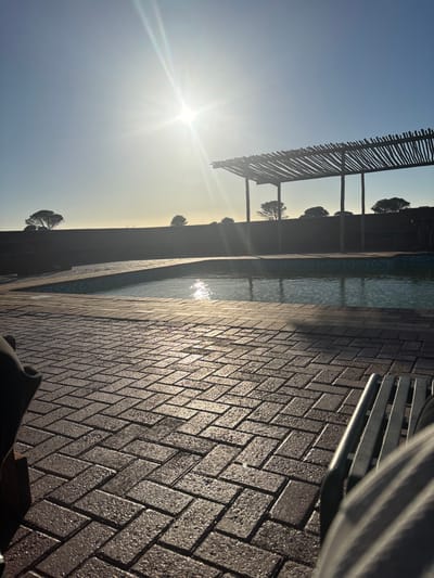 The image depicts an outdoor swimming pool area in Port Alfred, South Africa, at either sunrise or sunset. The low sun, positioned in the upper center-left, casts significant lens flare and long shadows.

In the immediate foreground, a patterned brick paver surface, appearing damp or reflective, extends across the bottom half of the frame. On the far left, a dark, indistinct object, possibly part of a chair or a person's limb, is partially visible. On the far right, the metal armrest and striped fabric (likely clothing) of a seated individual are present.

The midground features a swimming pool with clear water, reflecting the bright sun. Behind the pool stands a rustic pergola structure composed of unhewn wooden poles and horizontal branches, providing partial shade. Beyond the pool and pergola, a dark, low-lying wall or embankment stretches across the horizon. Sparse, rounded trees are silhouetted against the bright sky on the distant horizon. The sky itself is clear, transitioning from a deep blue at the top to a bright, hazy white and yellow around the sun. No specific actions or interactions are observable.