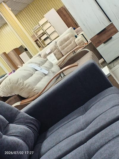 The image shows an interior view of what appears to be a furniture store. In the foreground, a dark gray upholstered couch or sofa is visible, with its cushioned surface filling the lower portion of the frame. Behind it, a lighter beige, tufted cushion is prominently displayed, with a curved wooden armrest or support extending across it. Further in the background, there are stacked furniture items, including what look like shelves and possibly cabinets or wardrobes. The overall impression is of a retail environment with various furniture pieces arranged for display. The image was taken on July 2, 2026, at 17:27, and the watermark indicates it was taken in the city of Chany, Russia.