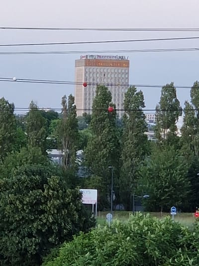 The image, taken in Fresnes, France, captures a view of a tall, cylindrical building with a patterned facade, identified as the Mercure hotel. The building's upper section features a sign that reads "Mercure". The building is surrounded by lush green trees, obscuring much of the lower half of the structure. In the foreground, dense foliage dominates the lower portion of the frame. Several power lines stretch across the upper part of the image, with a white sphere and a red sphere attached to them. A distant sign reading "NLYS" is partially visible through the trees. The sky is overcast, suggesting a cloudy day.