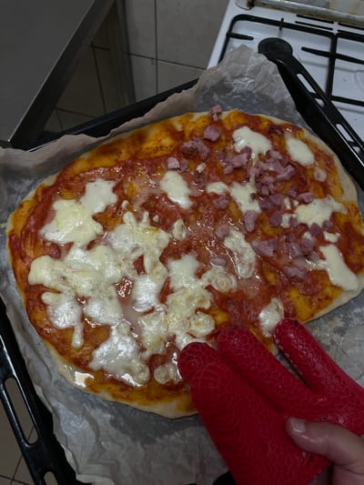 In Dumbrăvița, Romania, a freshly baked pizza rests on a black baking tray lined with parchment paper. The pizza, adorned with tomato sauce, melted mozzarella, and diced ham, appears to be close to completion, its cheese glistening. Two red silicone oven mitts are positioned near the pizza, with a hand, wearing one of the mitts, touching the edge of the tray, suggesting the pizza is either being removed from or placed into an oven. The background hints at a kitchen environment, with a stove visible.