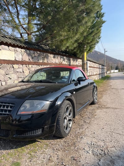 A black first-generation Audi TT convertible, featuring a contrasting red soft top, is parked on a dirt and gravel shoulder in Bryastovo, Bulgaria. The vehicle is angled slightly towards the left, with its front end facing the viewer and its left side parallel to an old stone wall constructed with irregular stones and horizontal wooden beams, topped with dark corrugated roofing. A large pine tree with vibrant green needles is situated behind and above the stone wall, casting partial shadows. A yellow-painted utility pole is partially visible behind the tree and alongside the wall. To the right of the car, a narrow, paved asphalt road curves gently into the distance, bordered by a dirt and grass verge. In the far background, a prominent, rocky hill or mountain stands under a clear, bright blue sky. No individuals or active interactions are visible in the scene. The lighting indicates a sunny day.